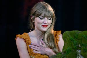 Taylor Swift smiling and placing a hand on her heart as she sits at a piano on the Eras Tour