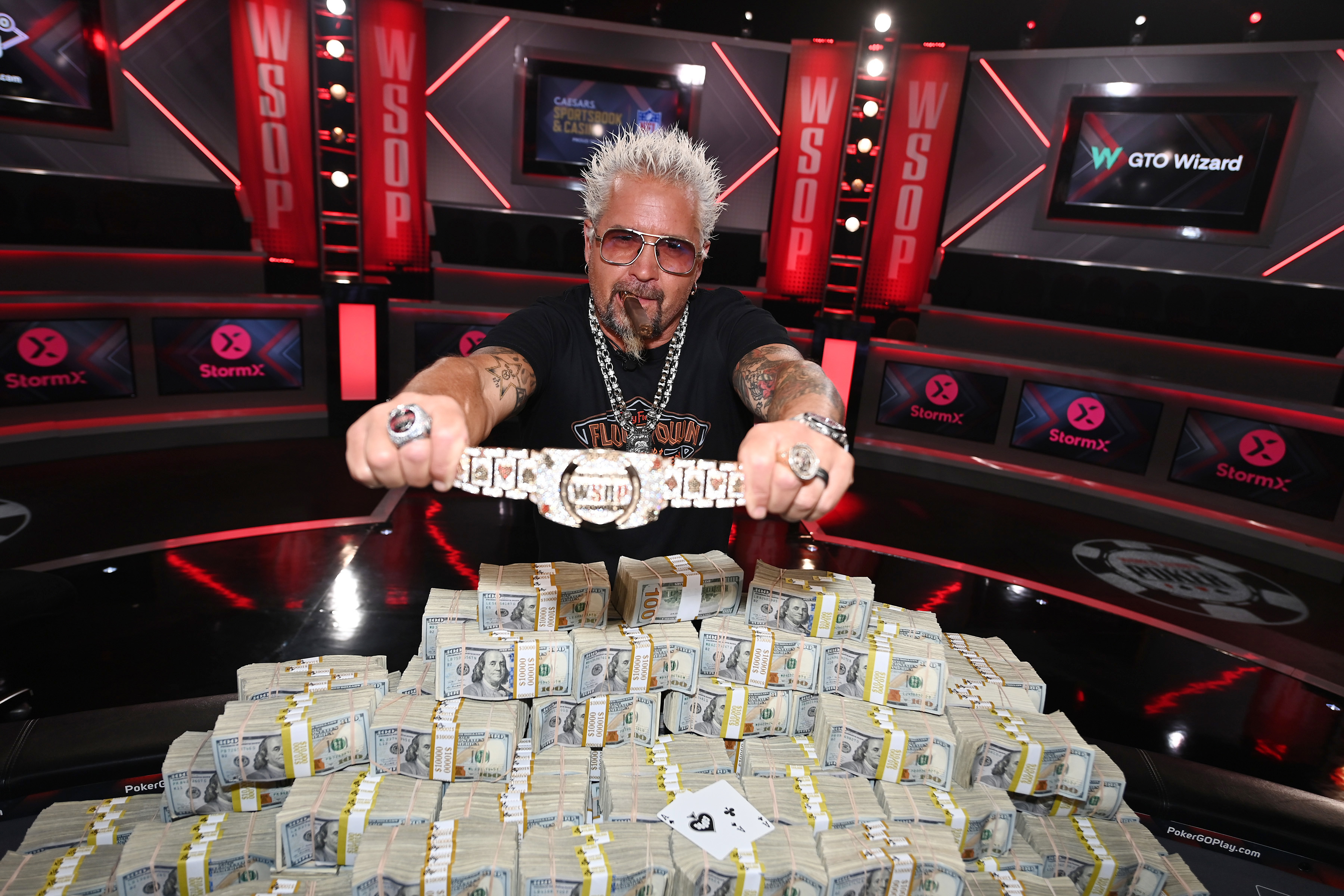 Guy Fieri holding up a winner&#x27;s belt as he stands behind a poker table full of cash with a cigar in his mouth