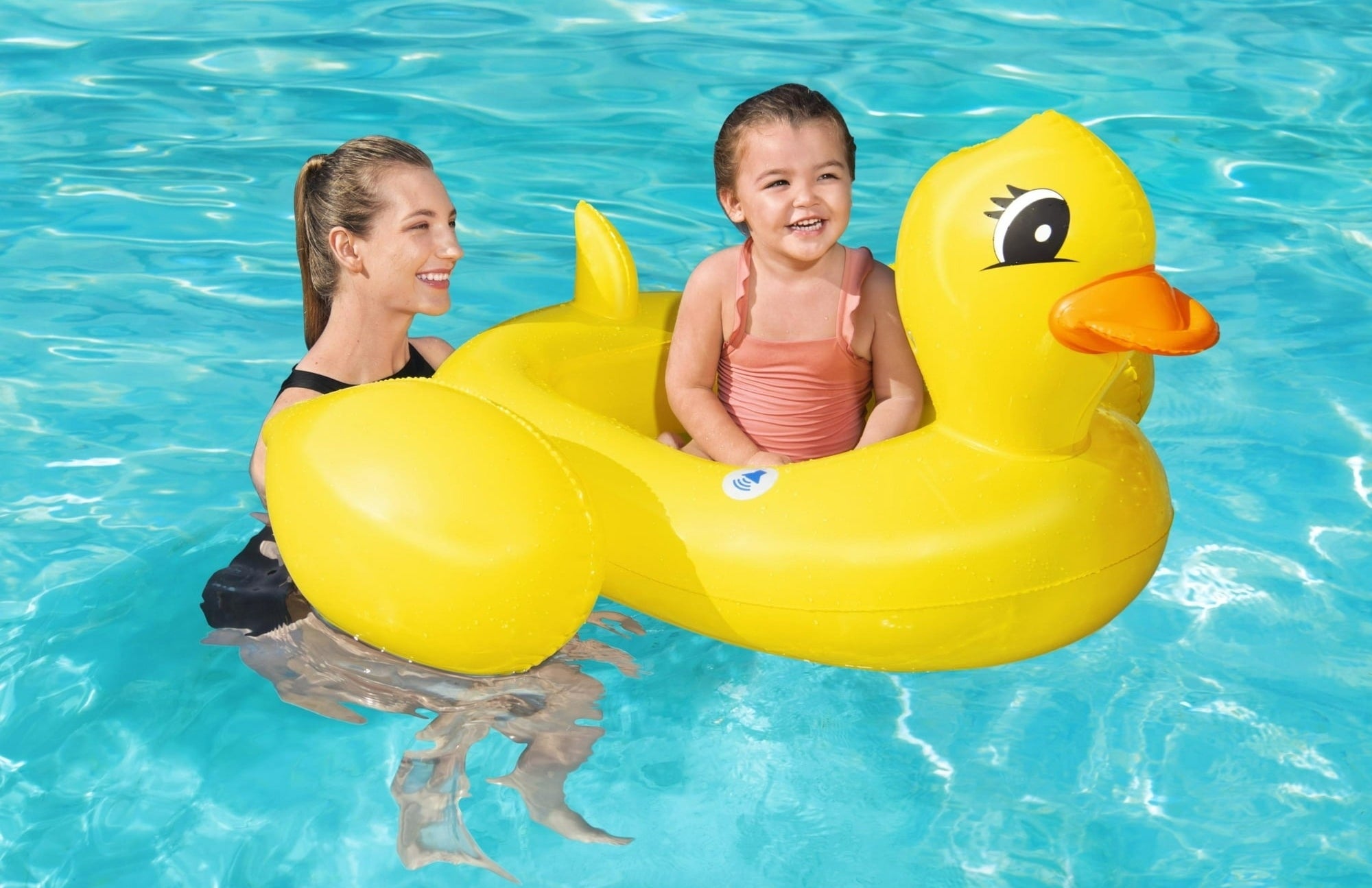 Smiling child in a yellow duck float is in a pool, accompanied by an adult. The scene exudes a playful and joyful atmosphere