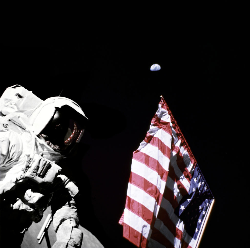 An astronaut with the US flag next to him and black expanse behind him