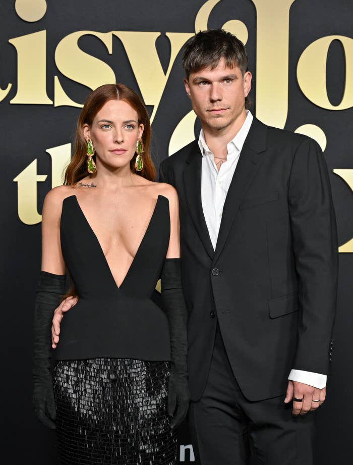 Riley Keough and Ben Smith-Petersen on the red carpet. Riley is wearing a dress with a deep v-cut and Ben is wearing a suit with no tie