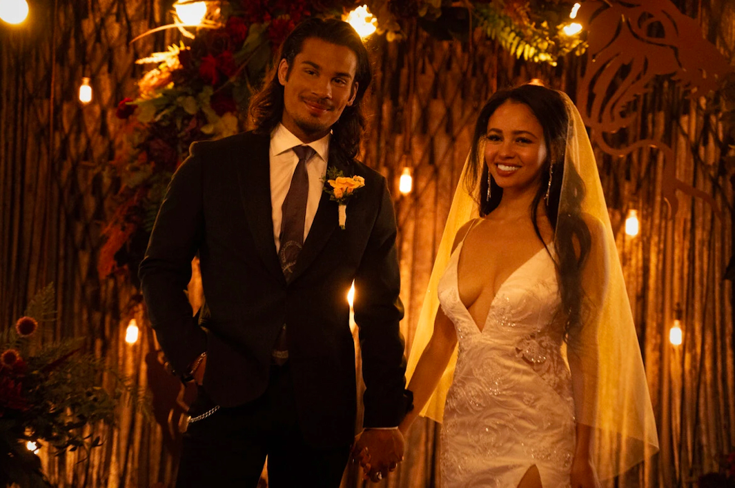 Fangs, a man with long, black hair holds hands with Toni, a woman with long, black hair. Fangs is wearing a black suit with a flower on his lapel, and Toni is wearing a white wedding dress and veil.