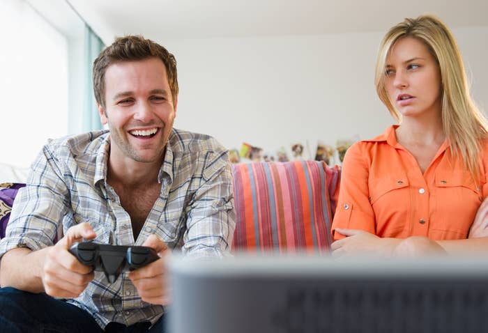 A man smiling and playing a video game, while a woman sits next to him, looking annoyed with her arms crossed