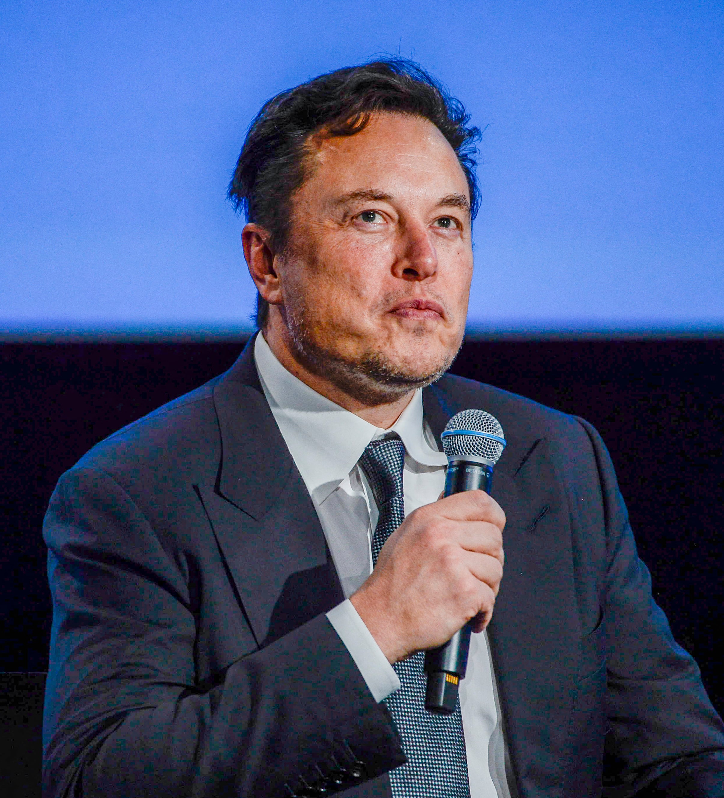 Close-up of Elon in a suit and holding a microphone