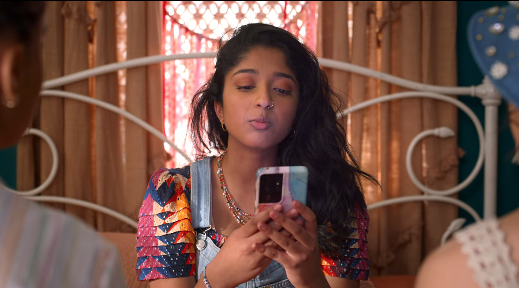 Devi from &quot;Never Have I Ever&quot; on Netflix sits on her bed, looks at her phone as if texting or on an app.