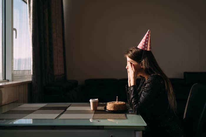 Cute thoughtful woman in party hat looking at festive cake with candle. Lonely woman freelancer celebrating birthday without friends. Quarantine and stay at home concept