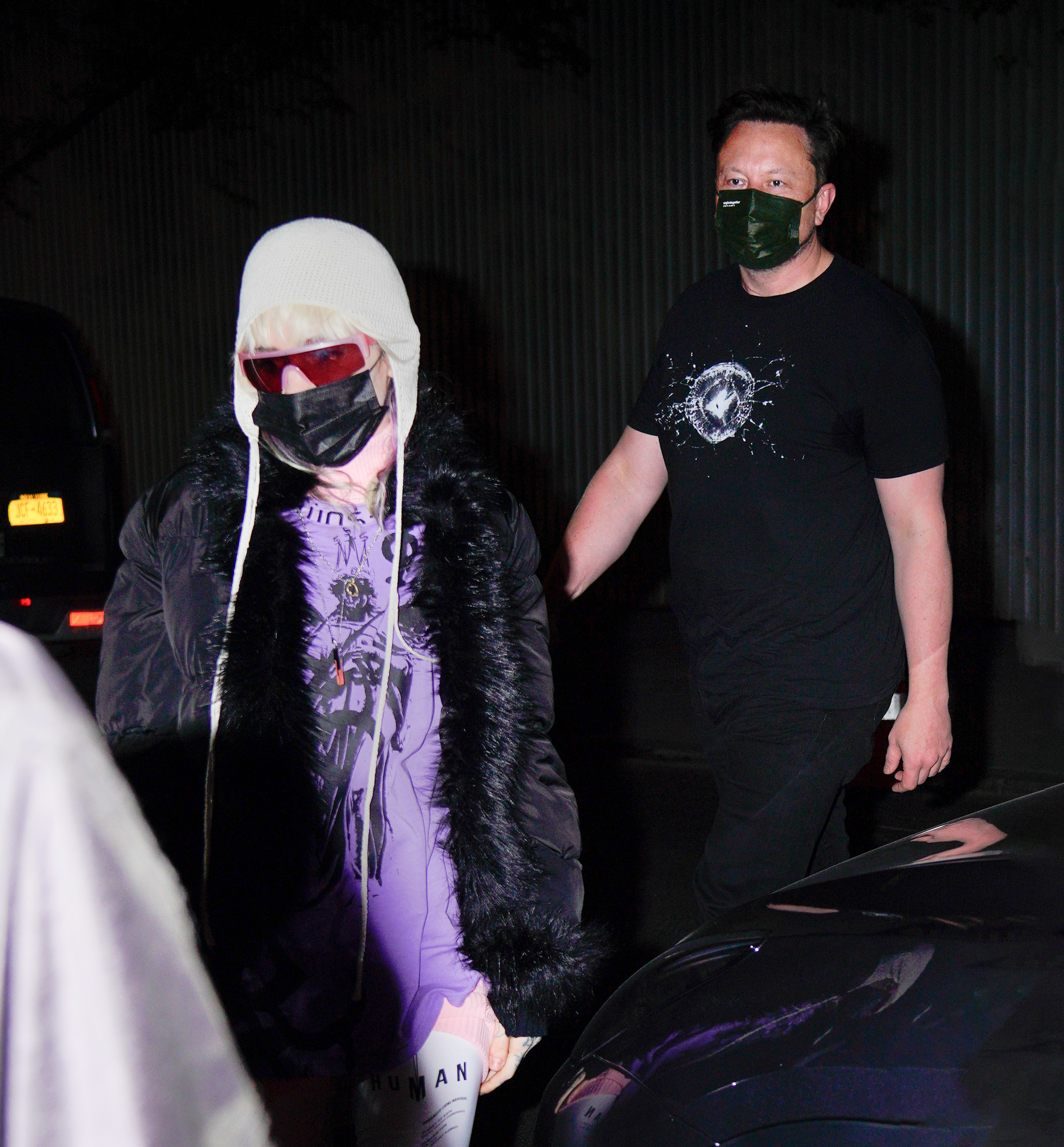 A closeup of Grimes and Elon wearing face masks and walking past a car