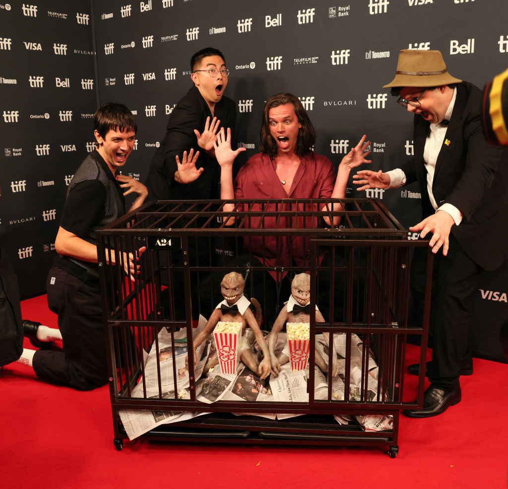 From left: Josh Sharp, Bowen Yang, Aaron Jackson, and Larry Charles pose on the TIFF red carpet. There is a little cage with scary looking dolls eating popcorn in front of them.