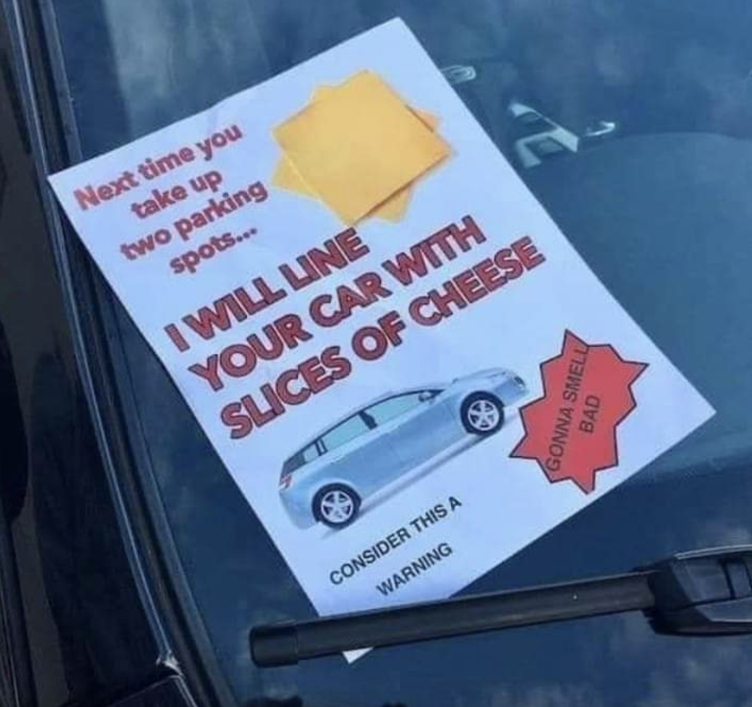 Sheet of paper with printed text tucked under a car windshield wiper: &quot;Next time you take up two parking spots, I will line your car with slices of cheese; consider this a warning — gonna smell bad&quot; with images of sliced American cheese and a car