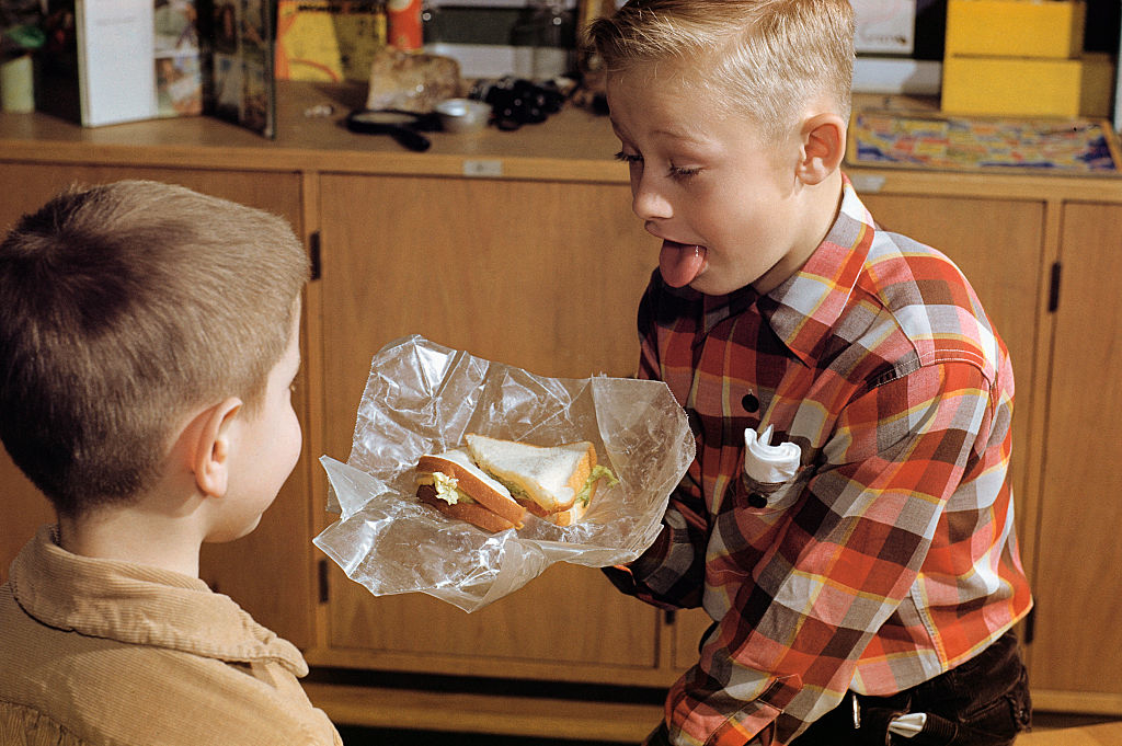 A kid showing another kid his sandwich with his tongue out