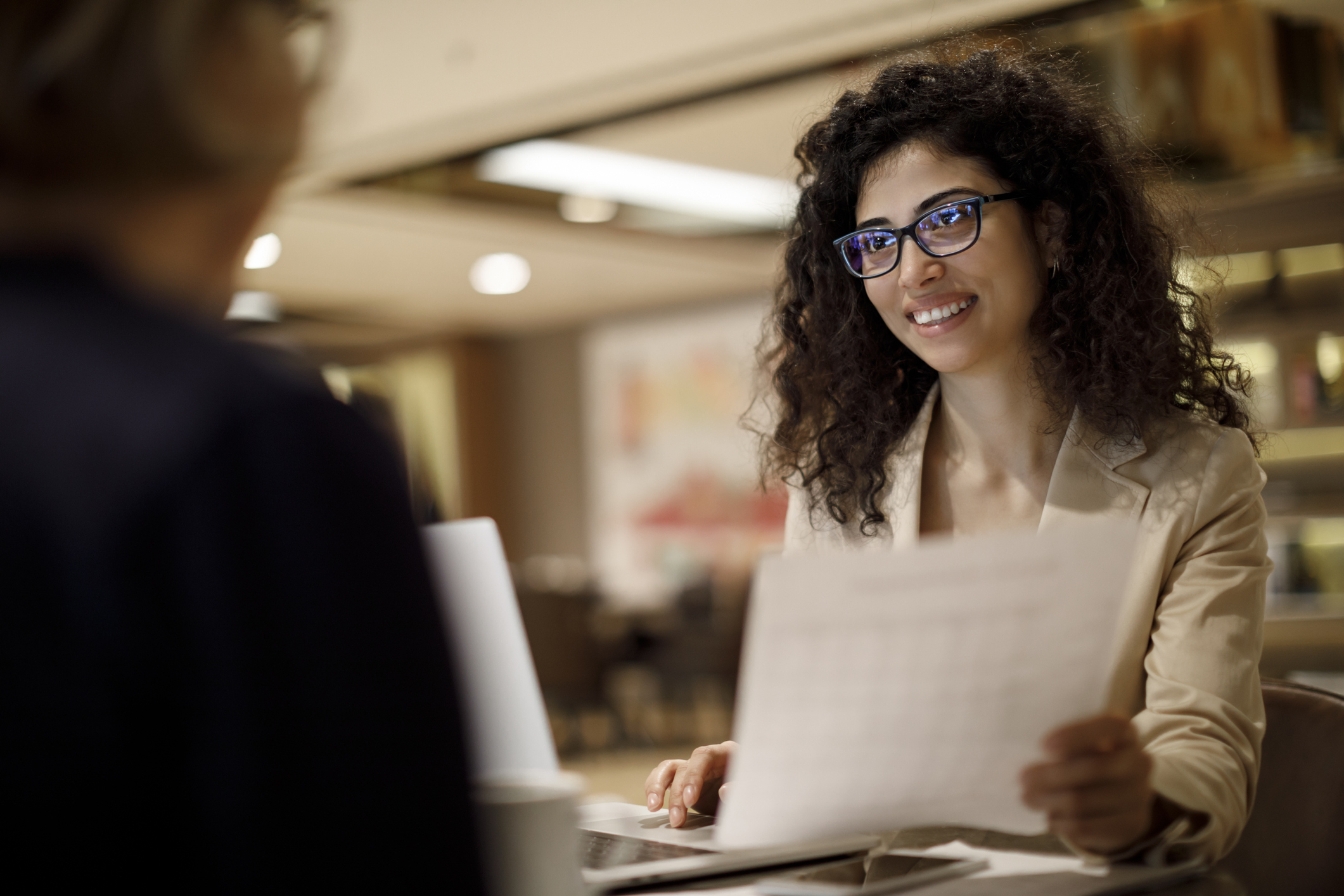 A smiling woman at a laptop talks to another person