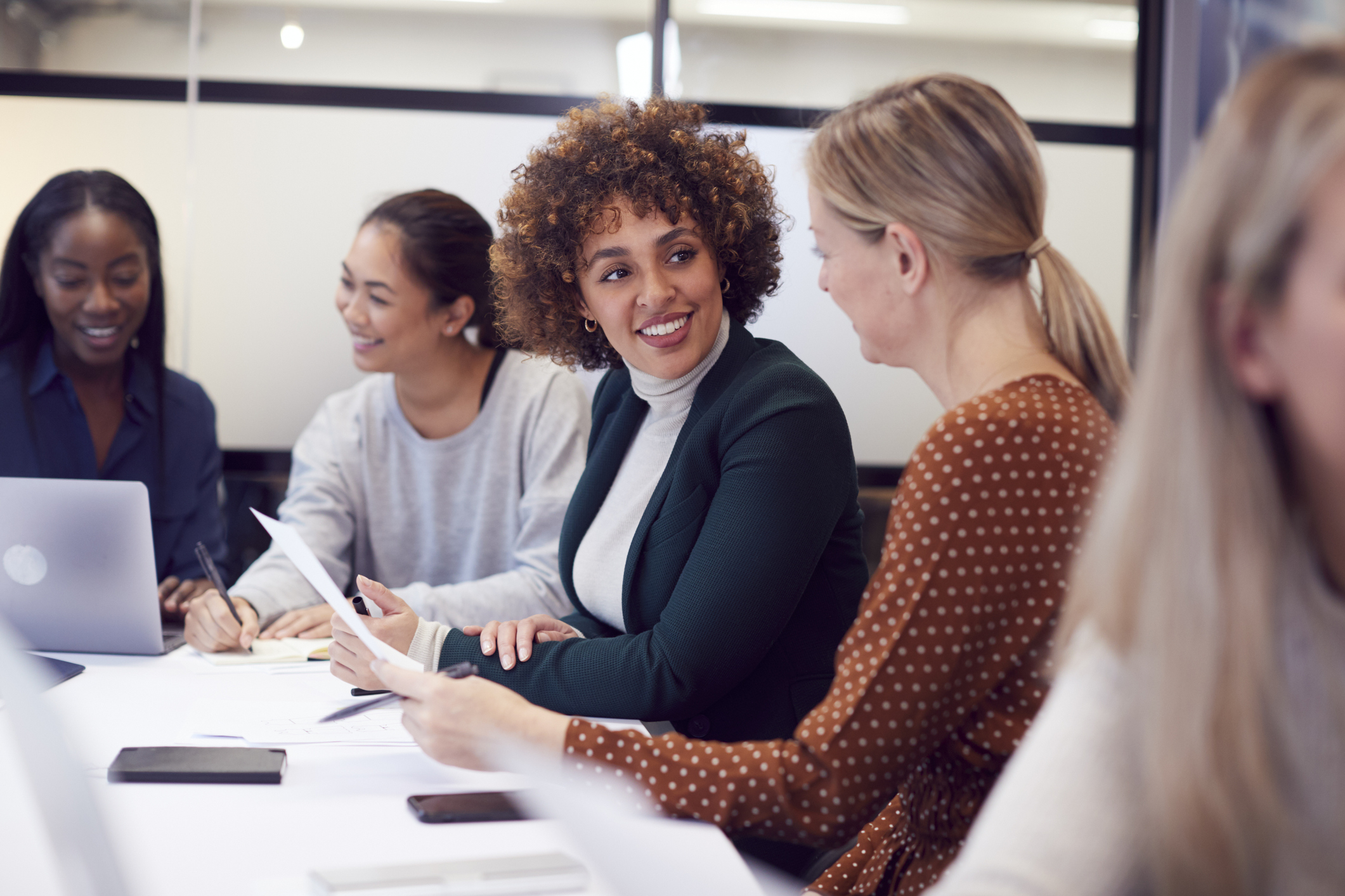 Smiling women at a table with laptops, papers, and phones