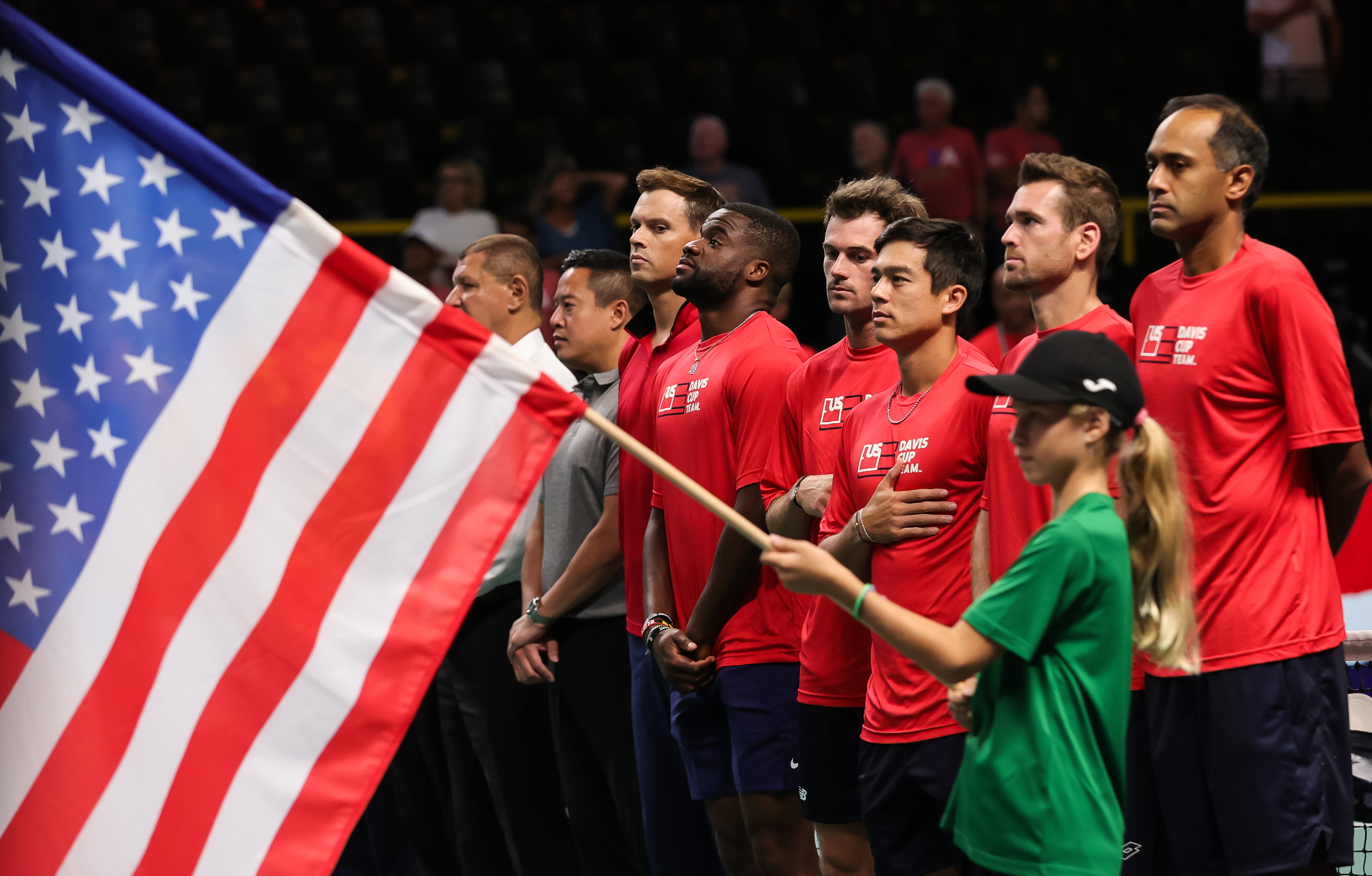Young girl holding a flag as athletes stand by her with their hands on their hearts