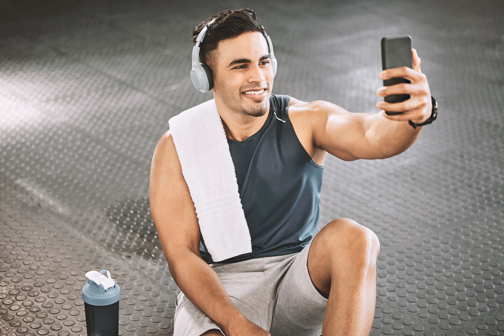 Man in a workout outfit with headphones and a towel on his shoulder taking a selfie