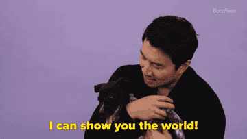 Simu Liu holds a puppy who licks his face. Text reads &quot;I can show you the world!&quot;