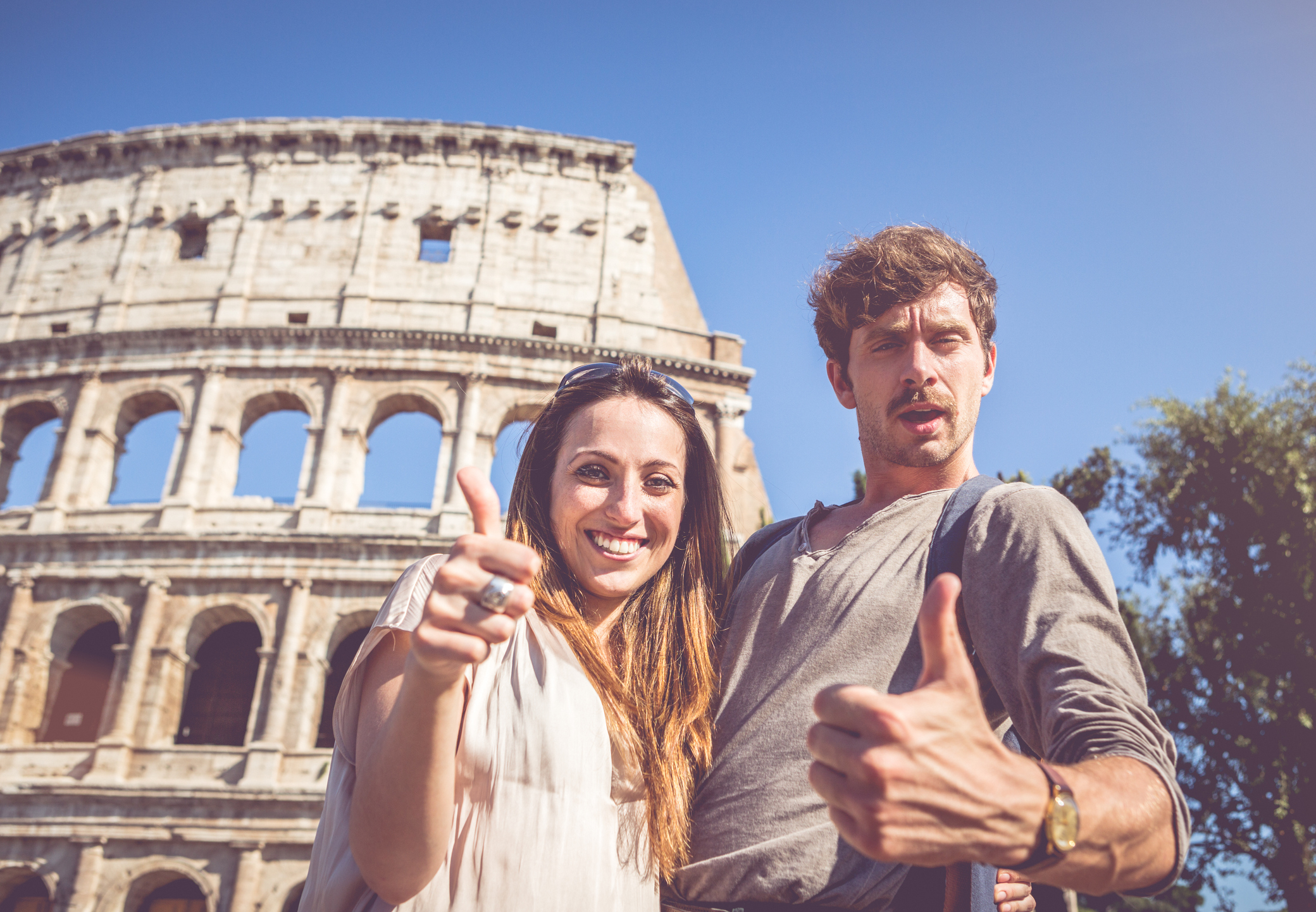 two tourists give a cheesy thumbs-up to the camera while posing in front of the Colosseum