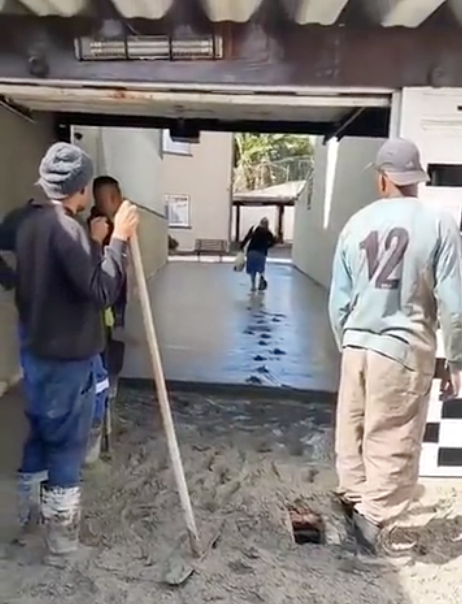 A group of workers watch as a woman trudges through clearly wet cement; she's halfway across the cement and has left a trail of footprints