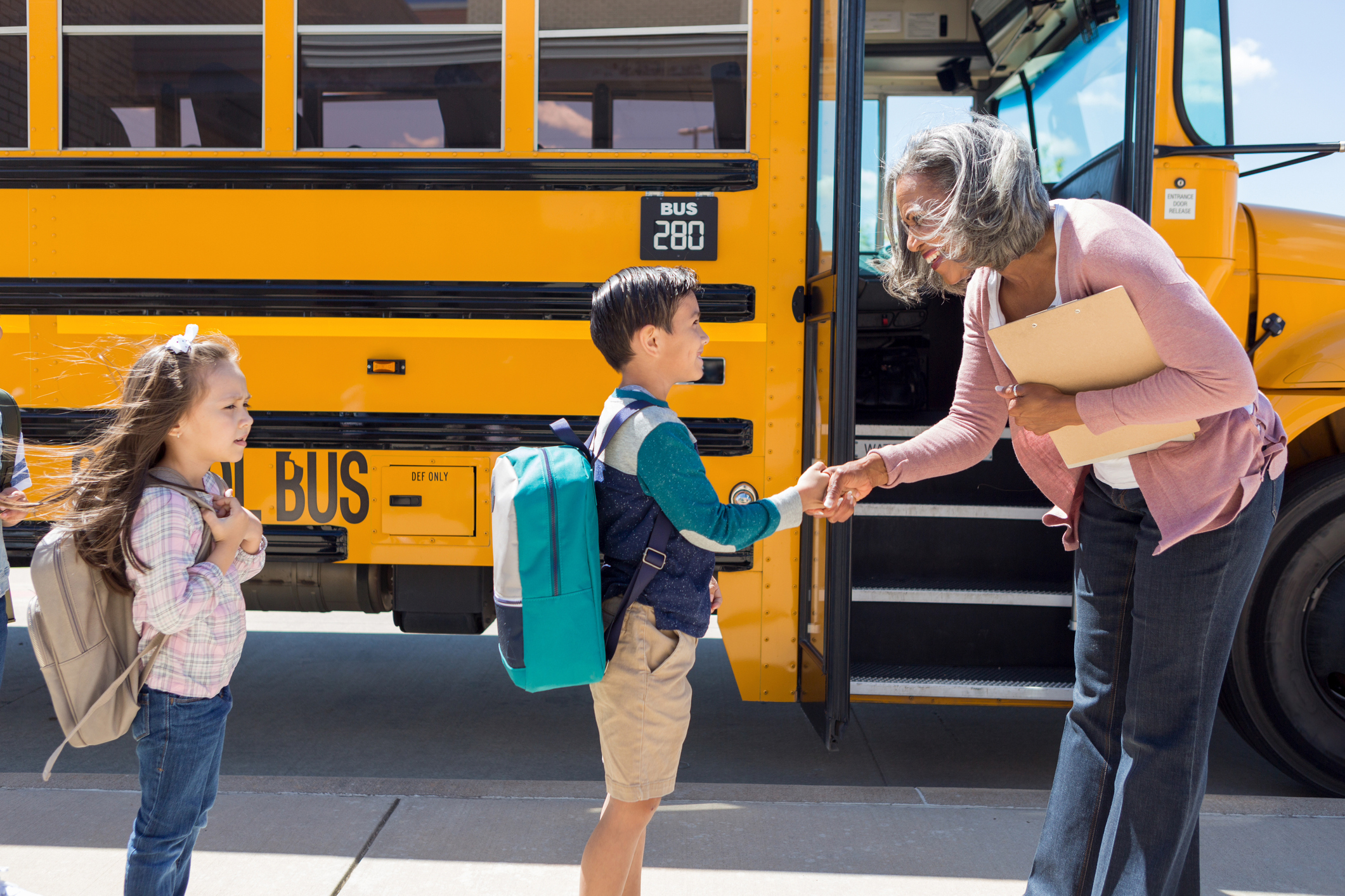 A young kid and a teacher are shaking hands in front of a school bus