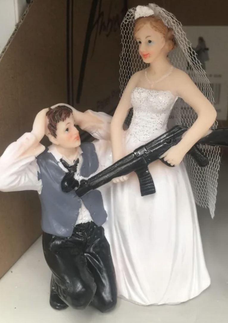 The topper shows the bride pointing a gun at the groom, who is kneeling with his hands on his head