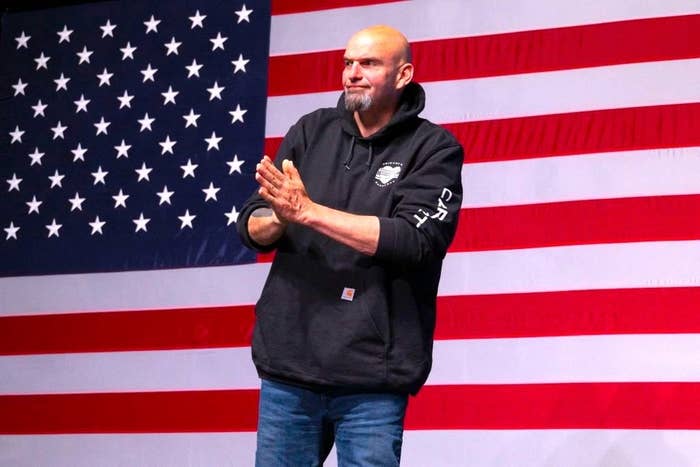 A Closeup of Sen. Fetterman onstage with a large U.S. flag behind him. Fetterman, who is quite tall, is wearing a hoodie and jeans