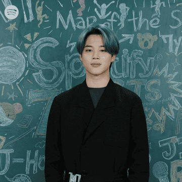 Jimin in front of a chalkboard background, cupping his face and smiling.