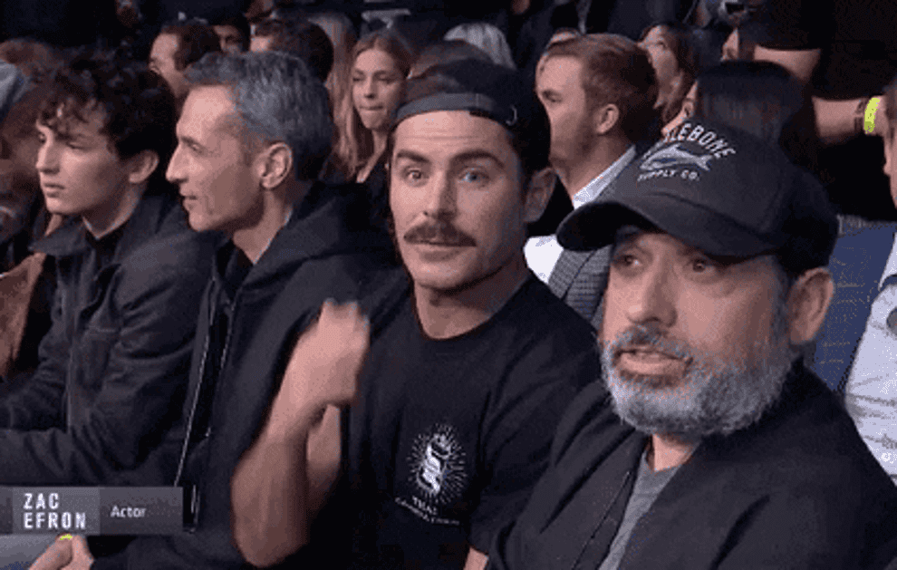 Zac Efron with a moustache waves and salutes and winks at the camera.