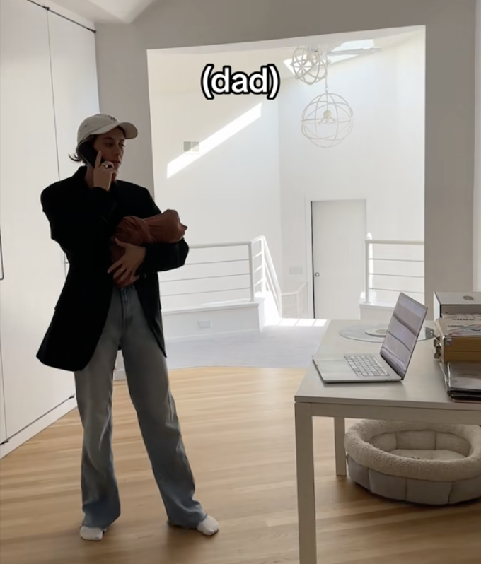 Dad on the phone carrying a baby with a laptop on a desk with caption "dad"