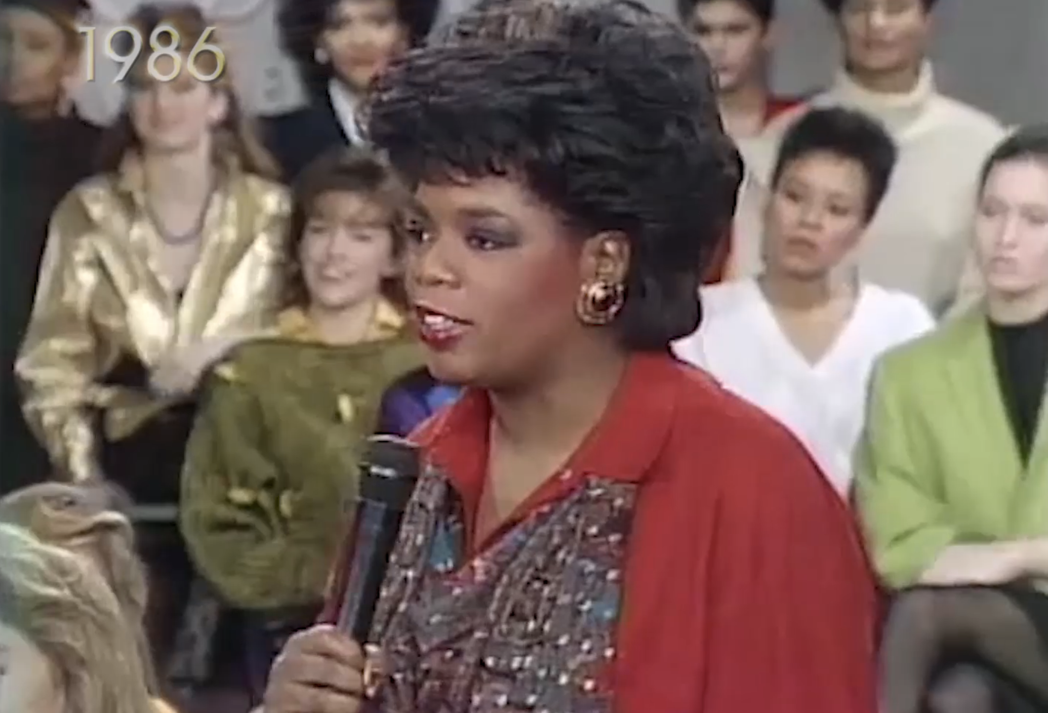 A closeup of Oprah holding a microphone with the audience behind her