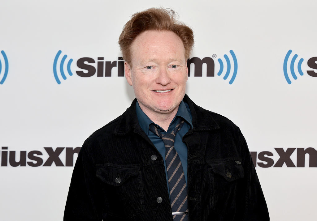Close-up of Conan smiling in a shirt, tie, and jacket at a media event