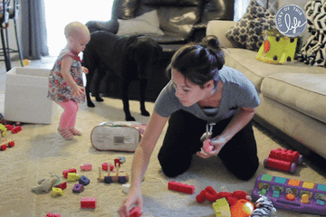 A woman picking up her baby&#x27;s toys while the baby dumps more on the floor