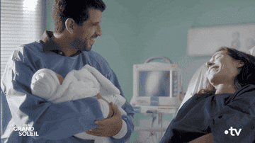 A man holding his newborn and smiling while the woman lays in the hospital bed and looks at him
