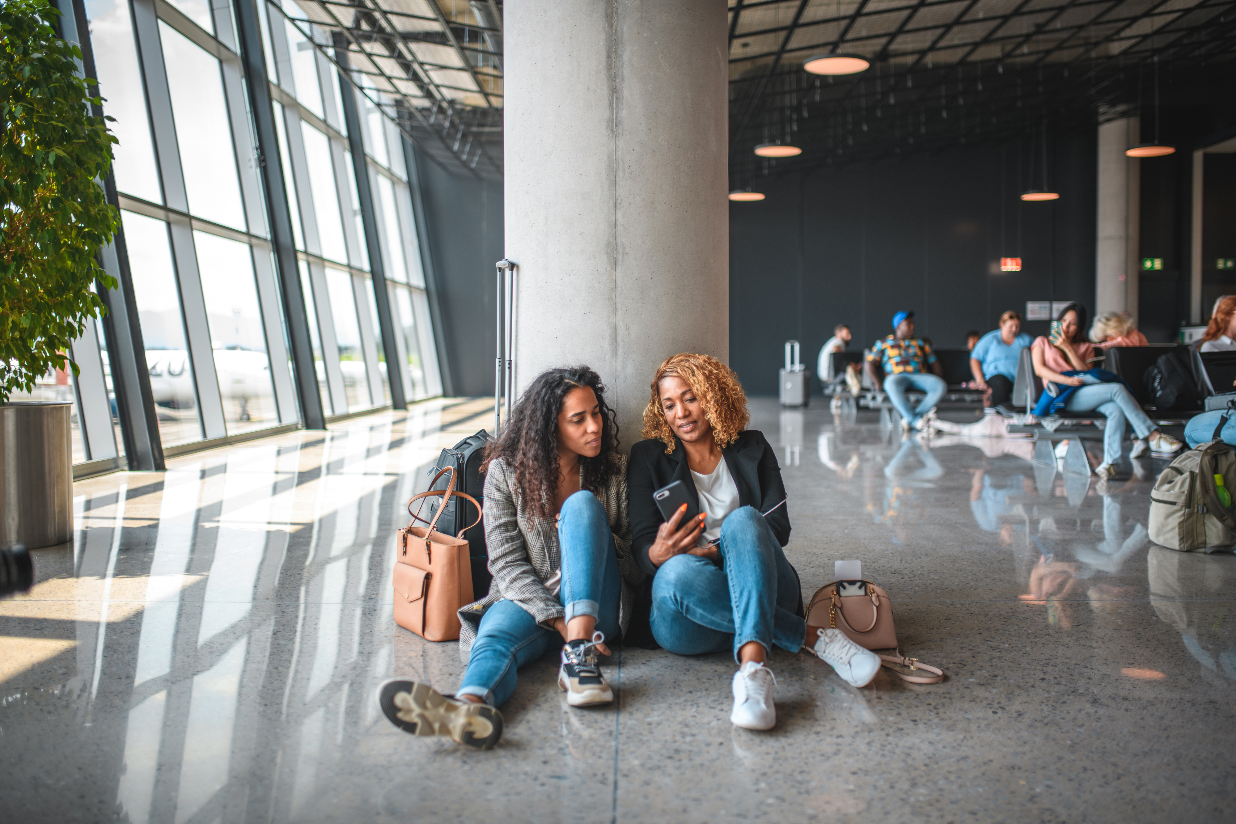 Hispanic mother and daughter sitting on the floor next to a window at the airport. Waiting for their flight using smartphone.
