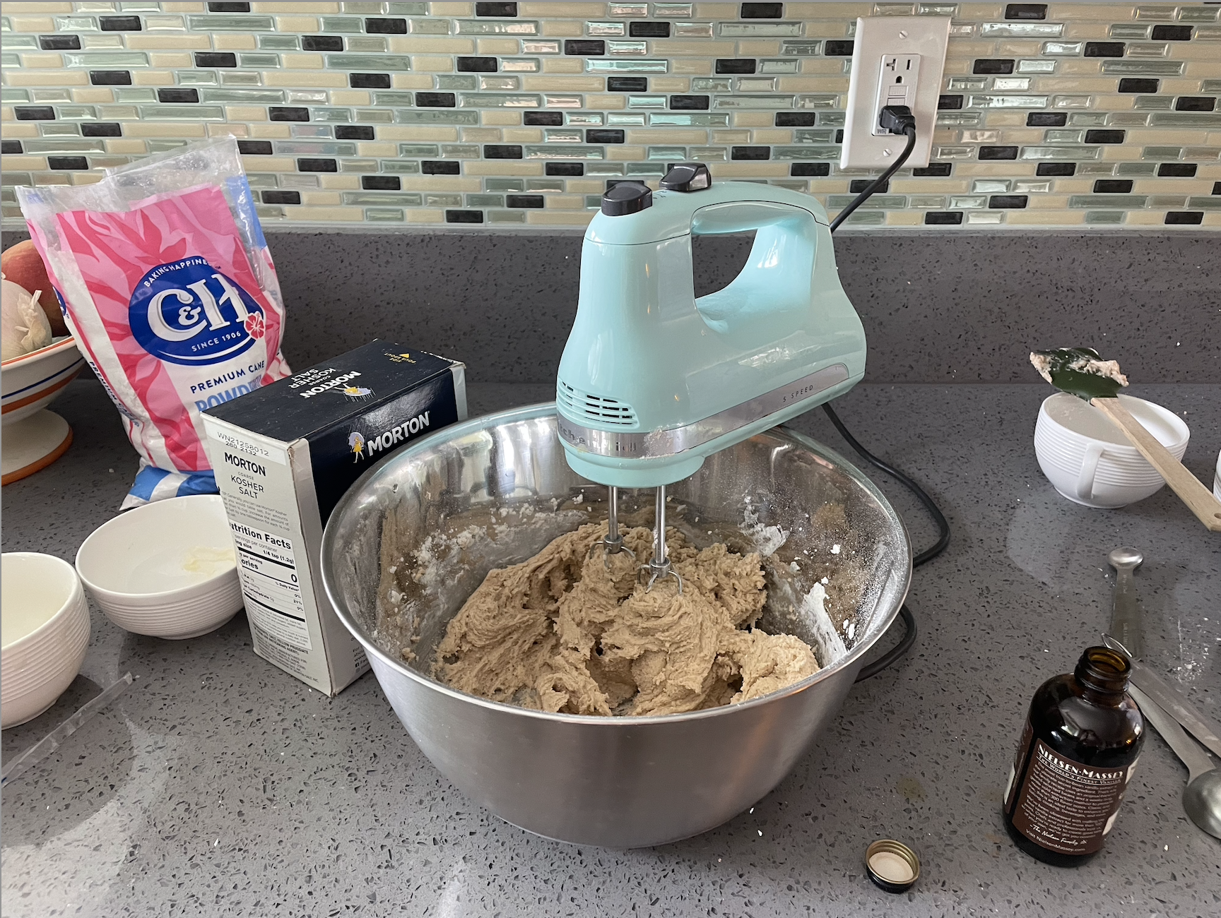 a hand mixer in a bowl of thicker cookie batter with flour added in; other ingredients like sugar, salt, and vanilla surround the bowl