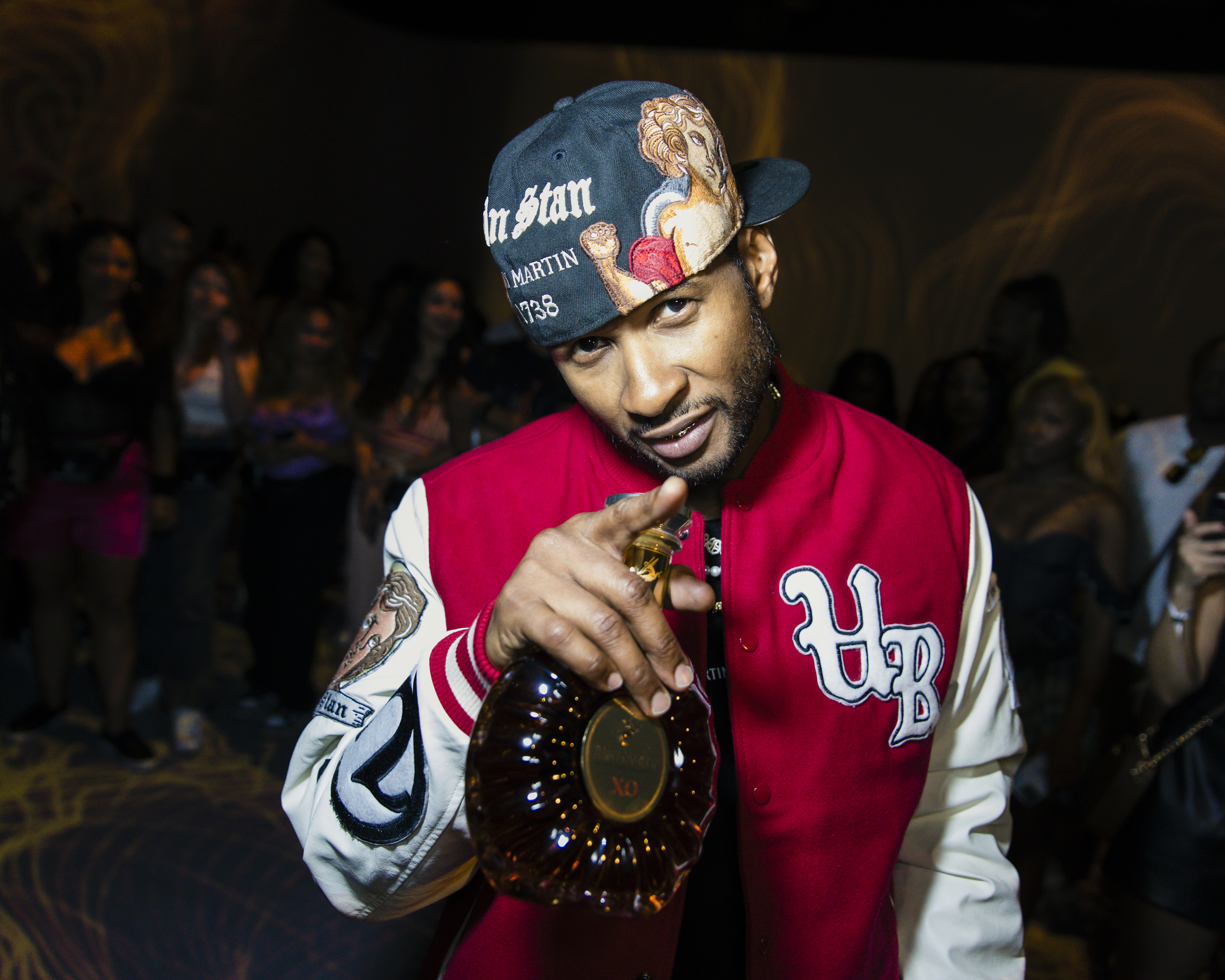 Usher at a party holding a bottle of liquor and pointing at the camera