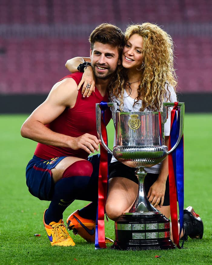 Shakira and Gerard smile as they kneel on a football pitch as they hold a trophy Gerard has just won