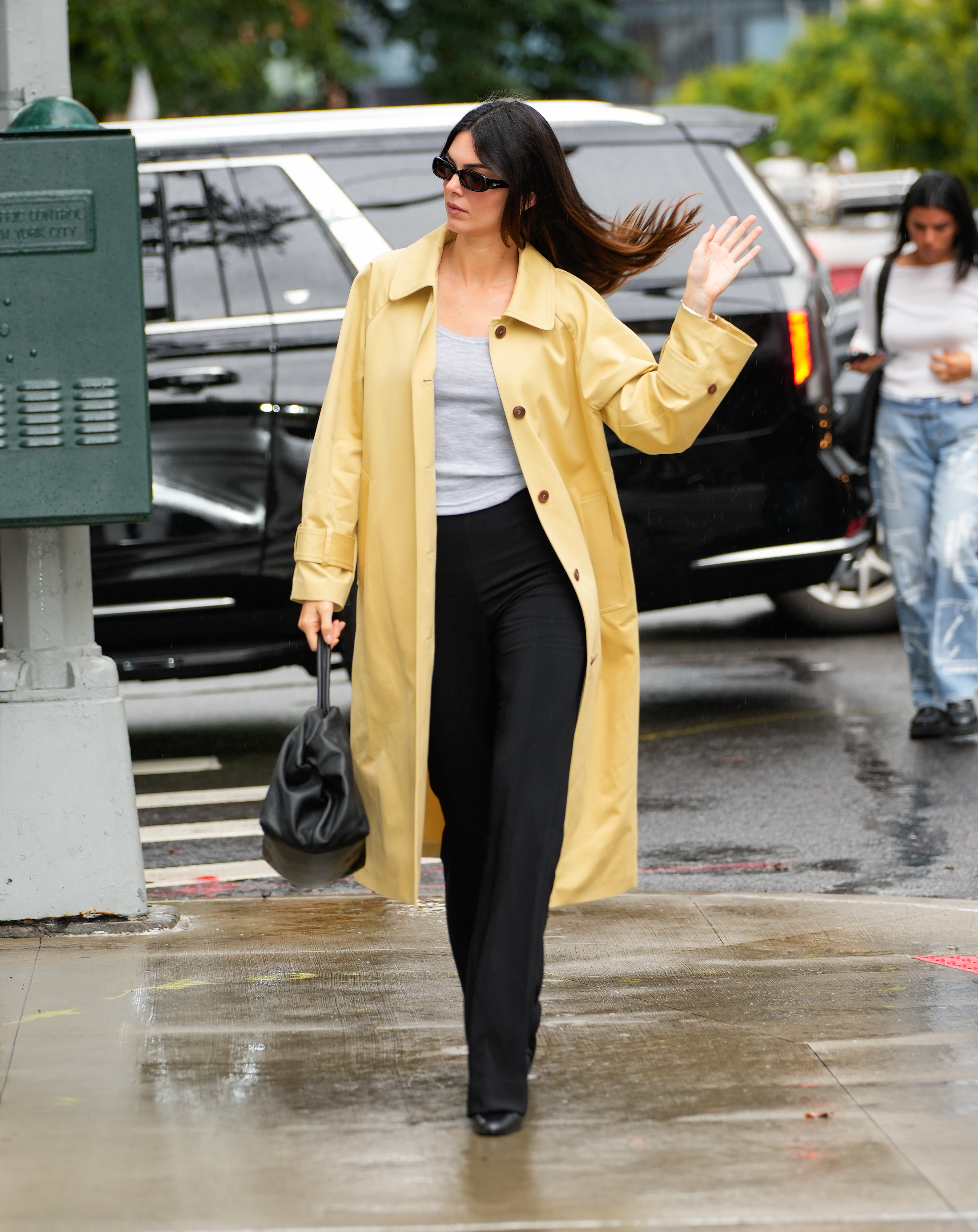 Close-up of Kendall in a trench coat and pants walking down the street and waving