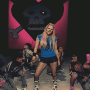 Avril Lavigne kicks her foot behind a pink skull background.