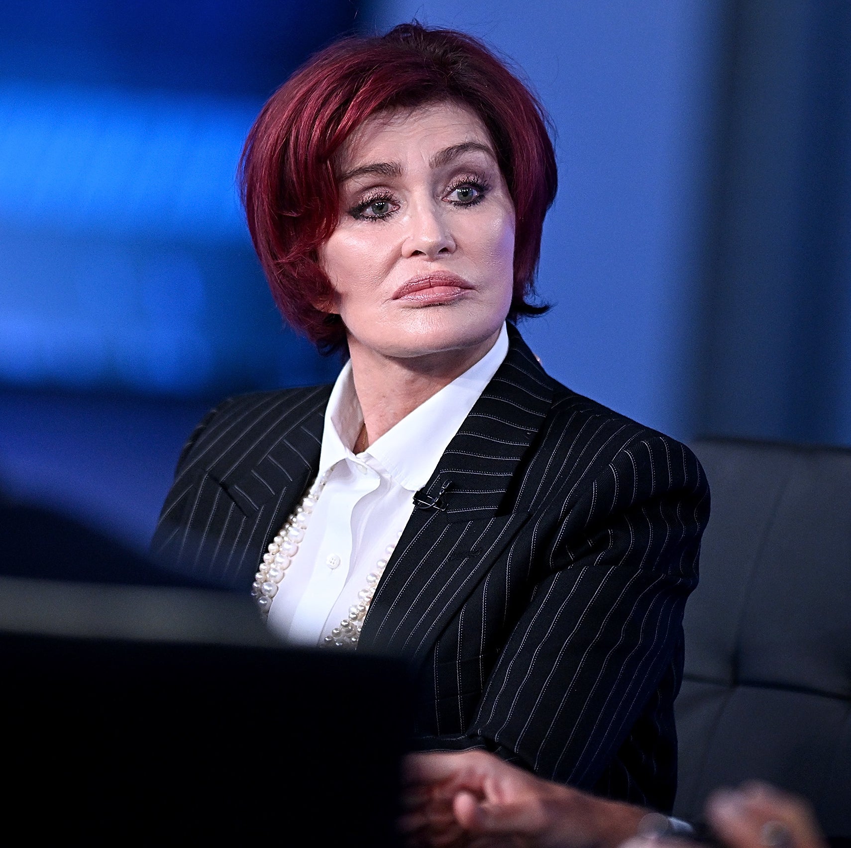 Close-up of Sharon wearing a pin-striped jacket, blouse, and a long pearl necklace