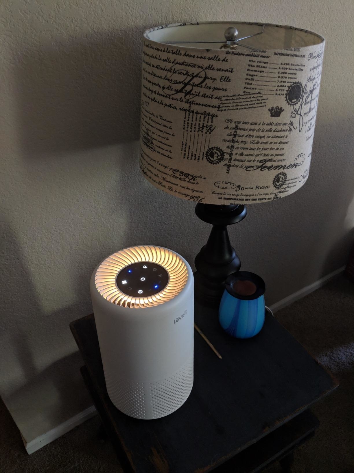 Reviewer image of the lit up air purifier on their nightstand next to a lamp