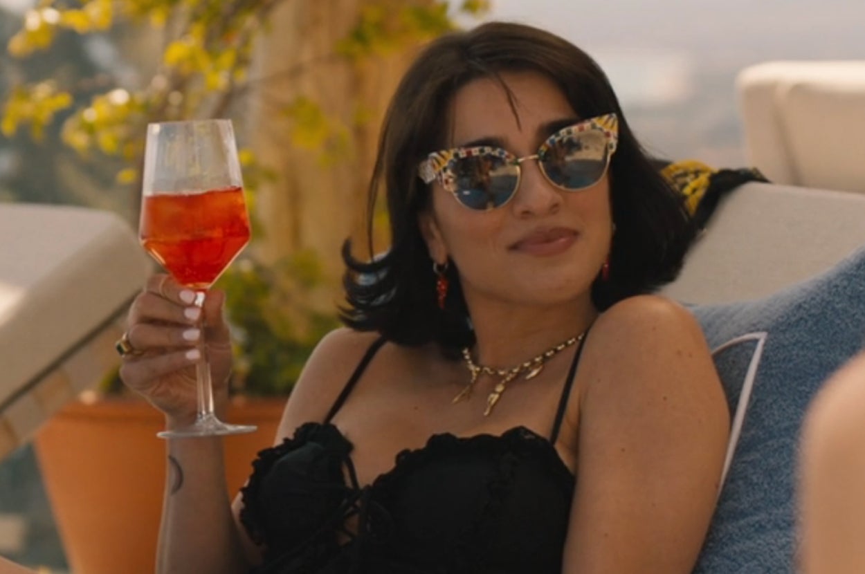 Lucia from The White Lotus sitting in a chaise by the pool and holding up a drink