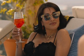 Lucia from The White Lotus sitting in a chaise by the pool and holding up a drink