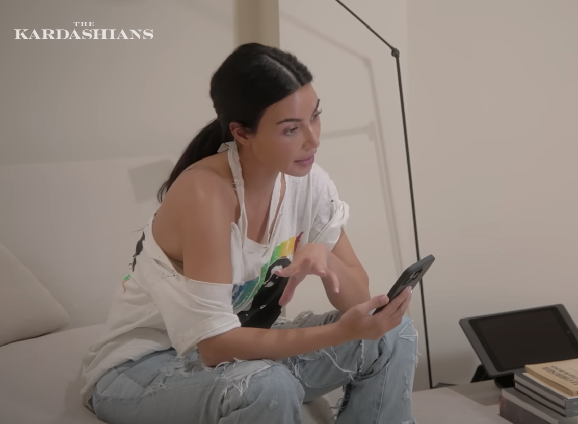 Kim sitting on a couch wearing frayed jeans and a torn T-shirt and looking at her phone