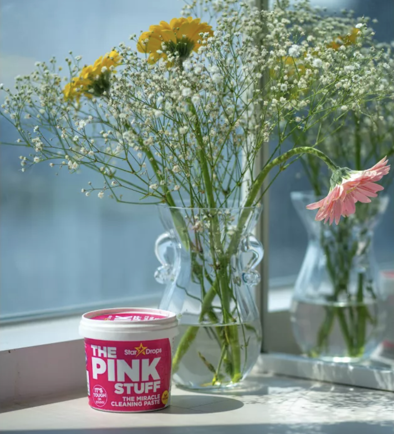 a container of The Pink Stuff on a windowsill with flowers