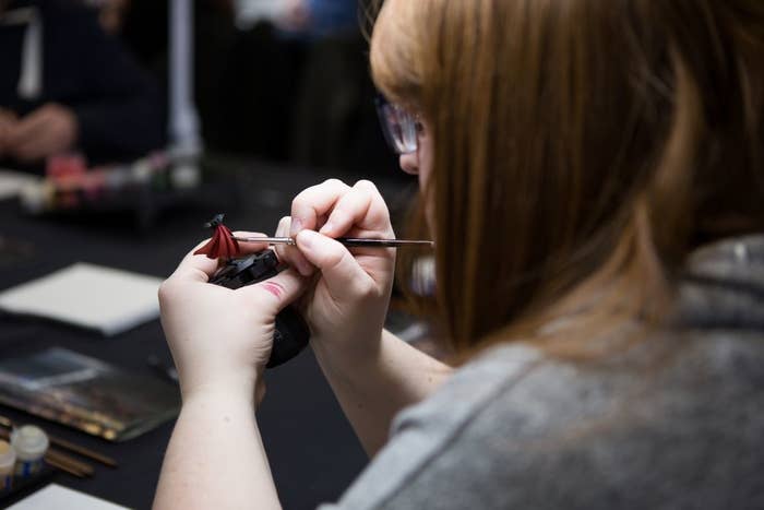 A woman in a grey t-shirt paints a Warhammer mini&#x27;s (I think it&#x27;s a Sister of Battle) robes red