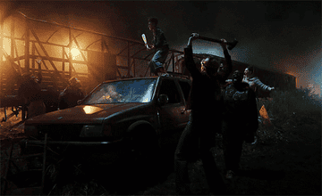 Girls hold bats and jump on top of a car in a junk yard.