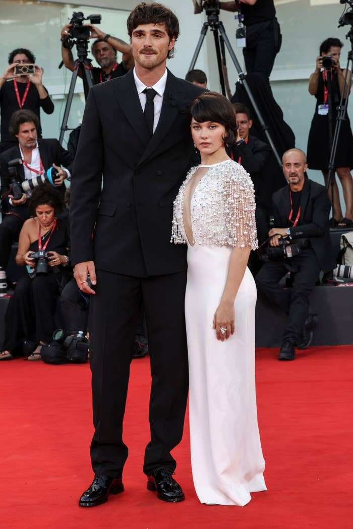 Closeup of Jacob Elordi and a much shorter woman posing on the red carpet with photographers in the background