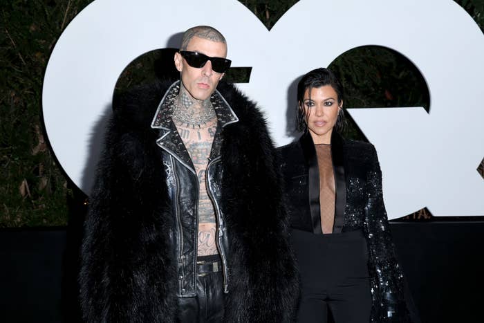 Travis Barker and Kourtney Kardashian stand side by side for photos at a GQ event. Travis is wearing an open coat with no shirt and Kourtney is wearing a sheer blouse and pants