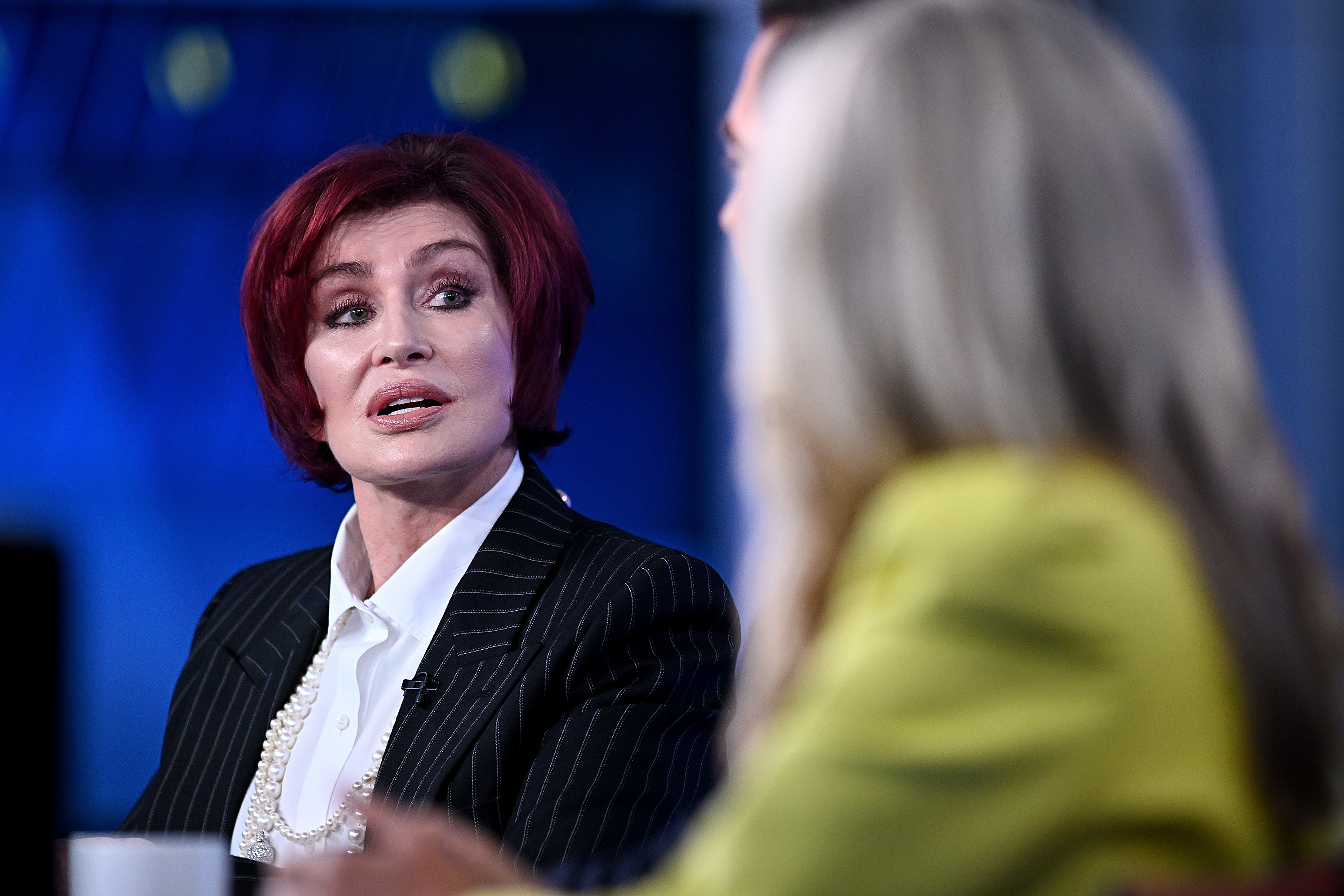 Close-up of Sharon wearing a suit jacket and pearls as she speaks to another person onstage