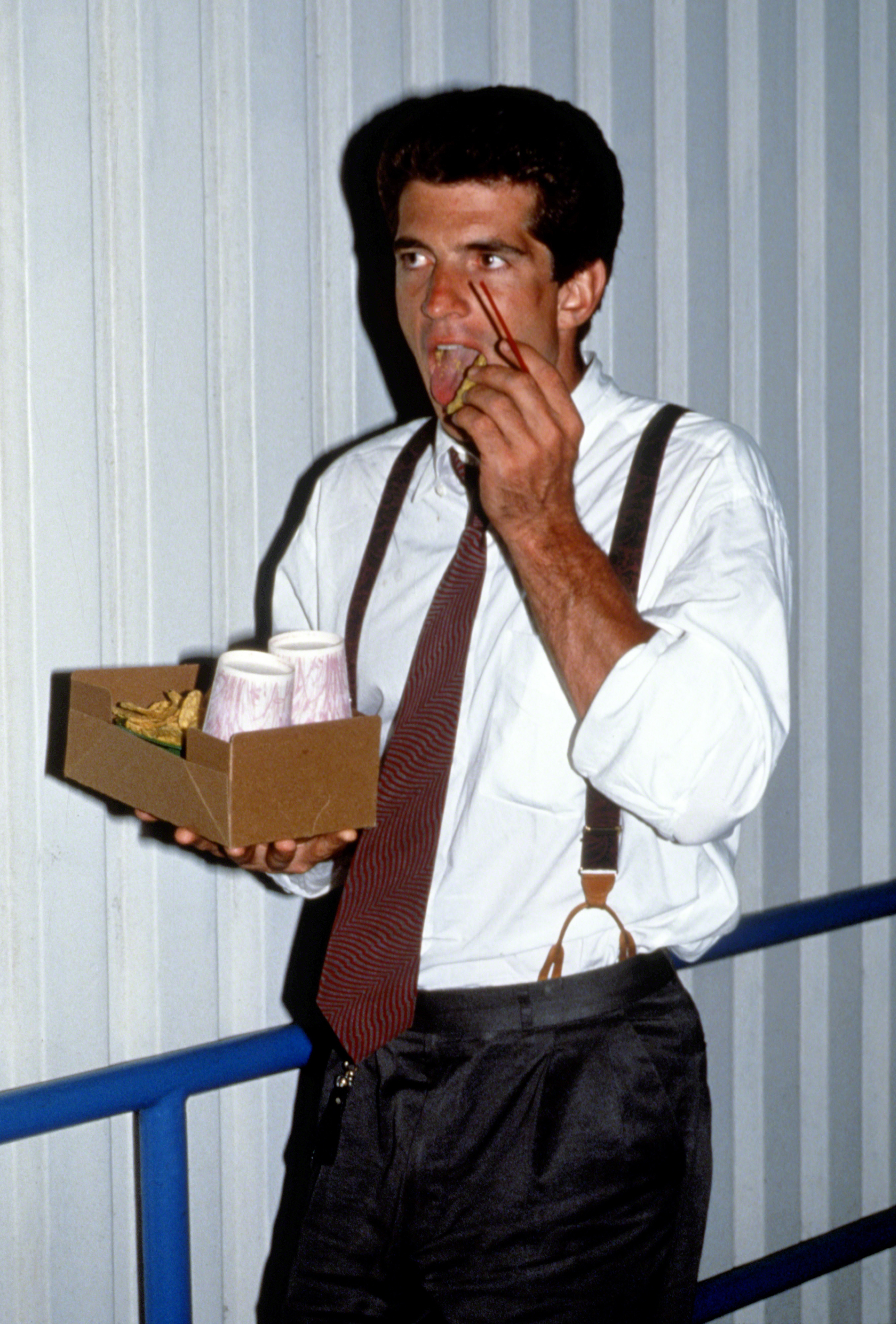 John F Kennedy Jr. walk as he holds a small paper basket of food and munches on a french fry; he&#x27;s wearing a dress shirt, tie, and suspenders