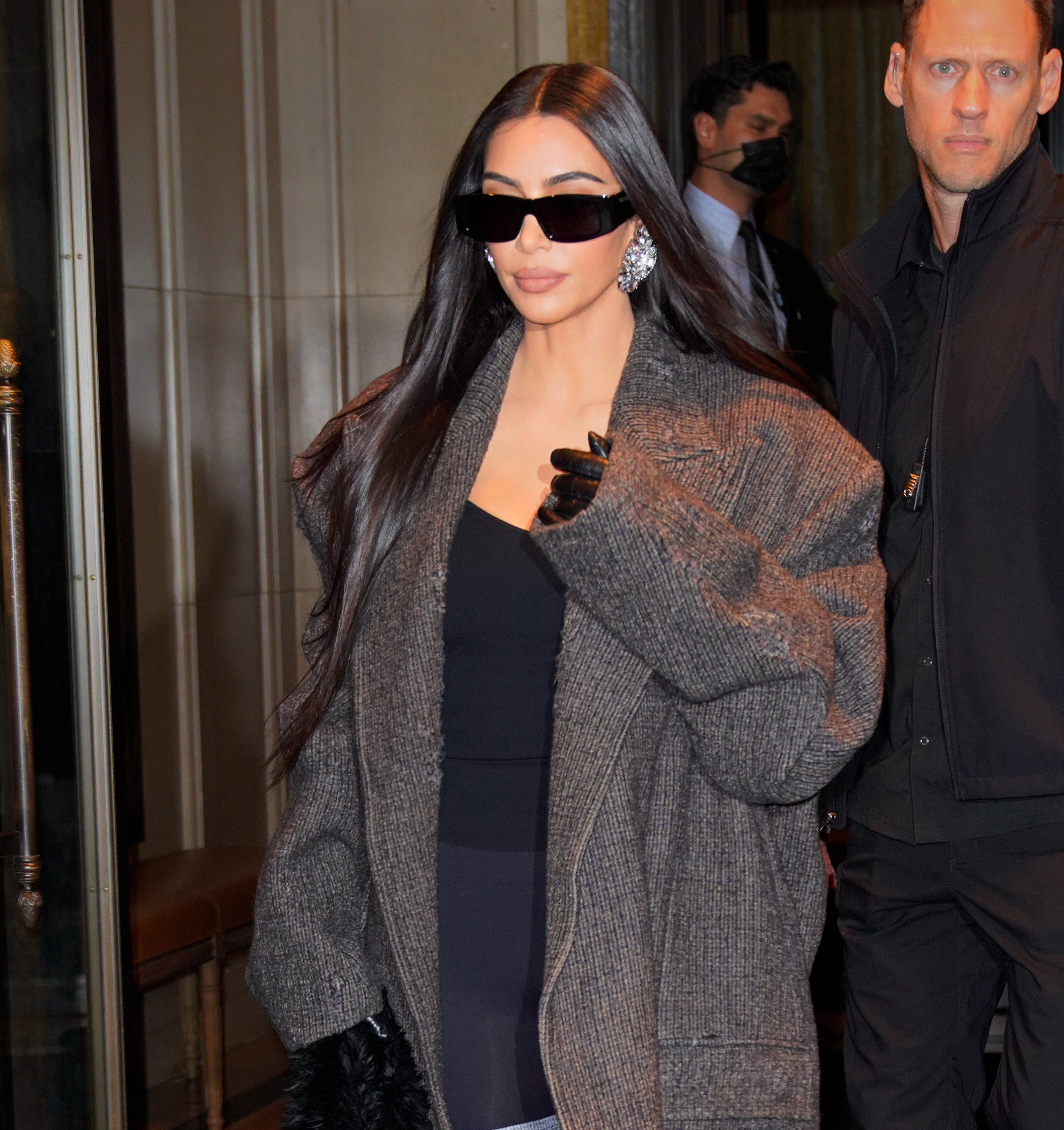 Kim walking out of a building wearing a large coat and sunglasses